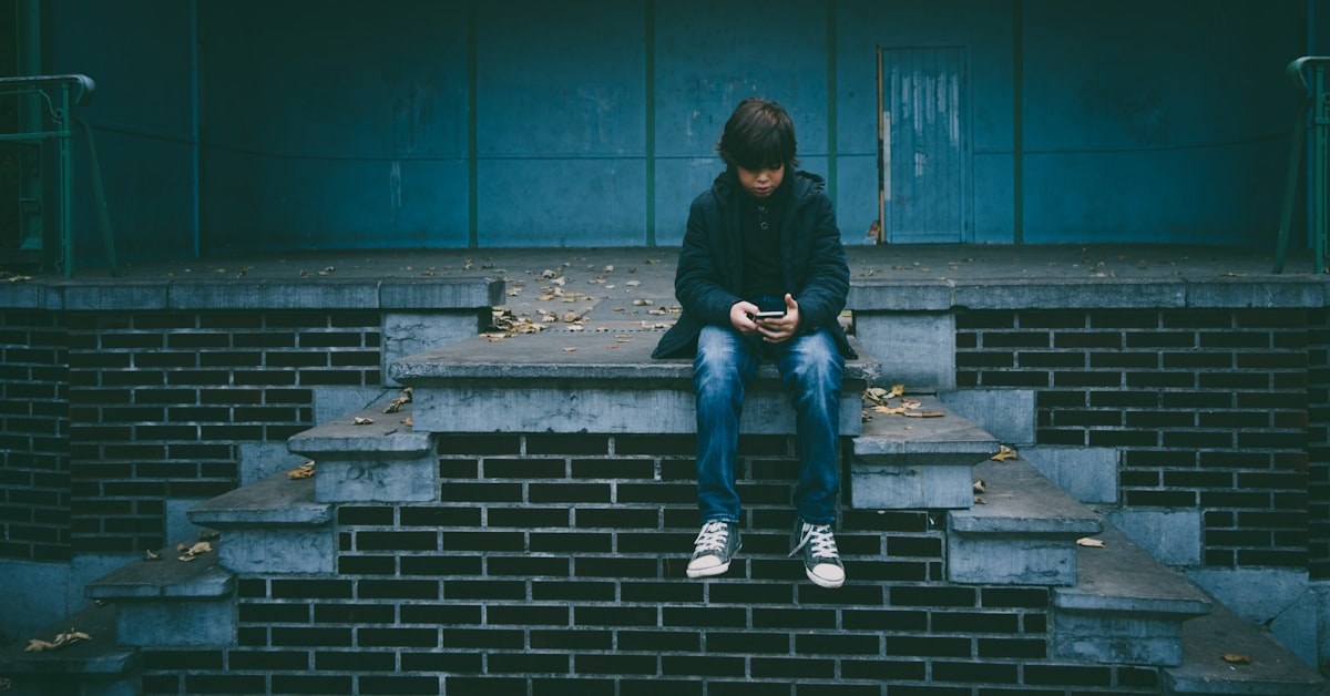 Jovem sentado olhando para o celular com expressão pensativa, representando o coração da geração digital por trás da tela