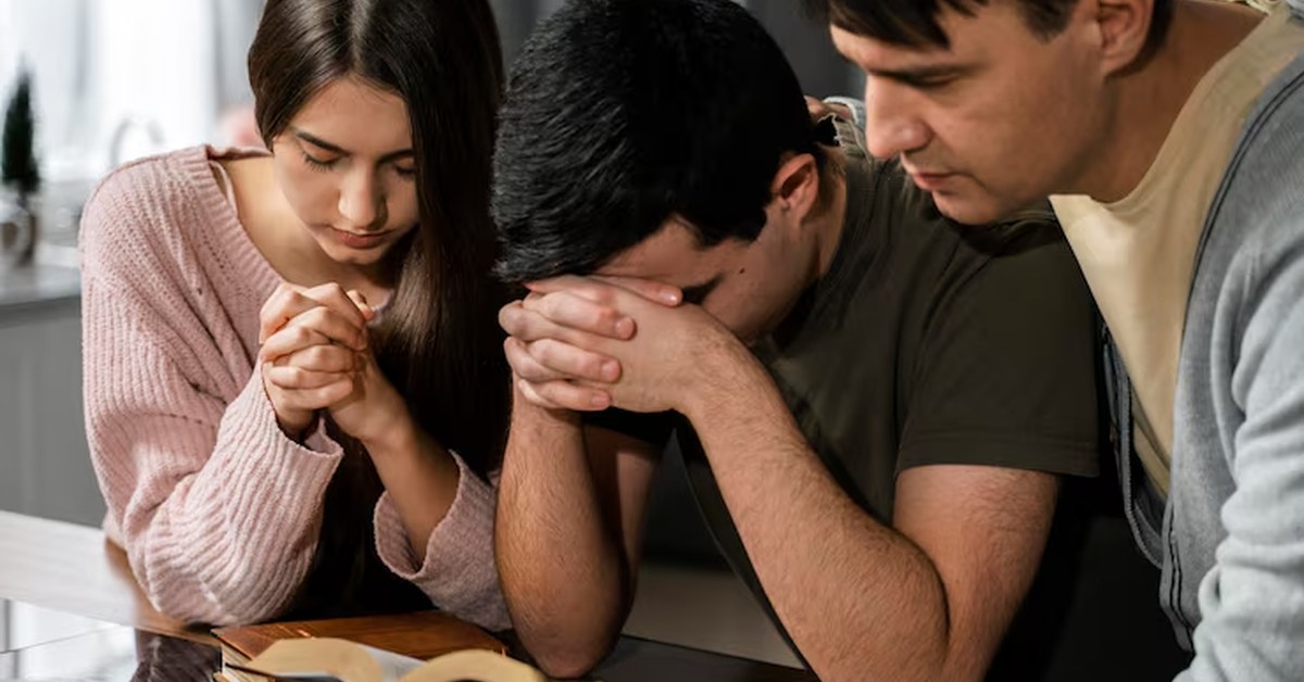 Adolescente e família orando juntos com a Bíblia aberta sobre a mesa, representando o momento devocional