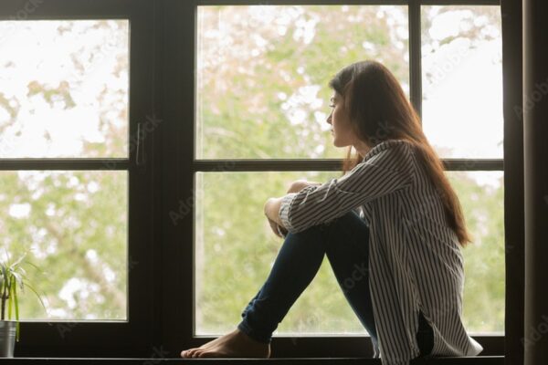 Adolescente sentado sozinho olhando pela janela, representando o isolamento sofrido por vítimas de bullying na infância